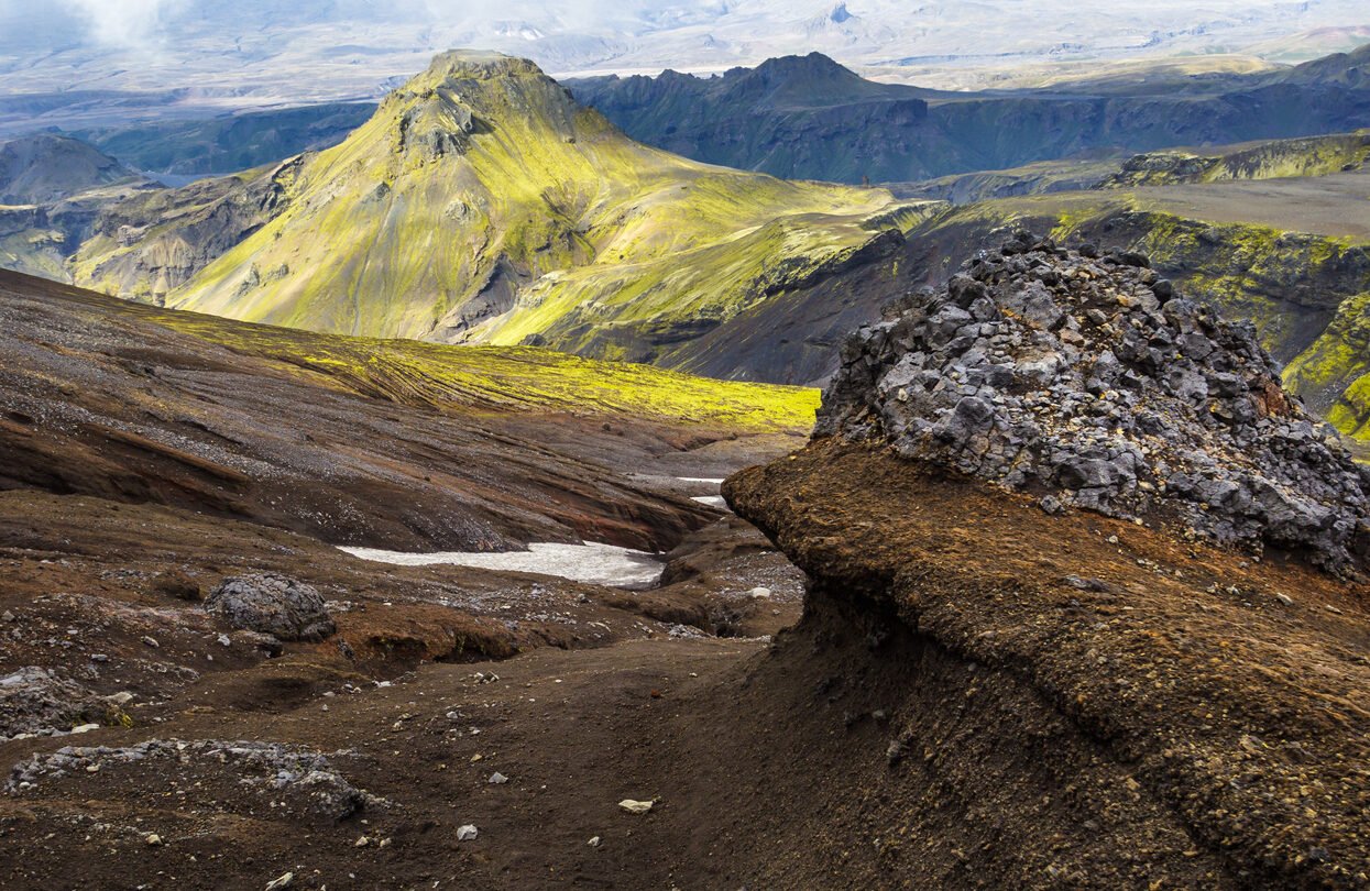 Hiking on Fimmvorduhals in summer: View over a landscape of bizarre lava formations, left over snow, moss covered mountains and the beautiful valley of Thorsmork in the distance.