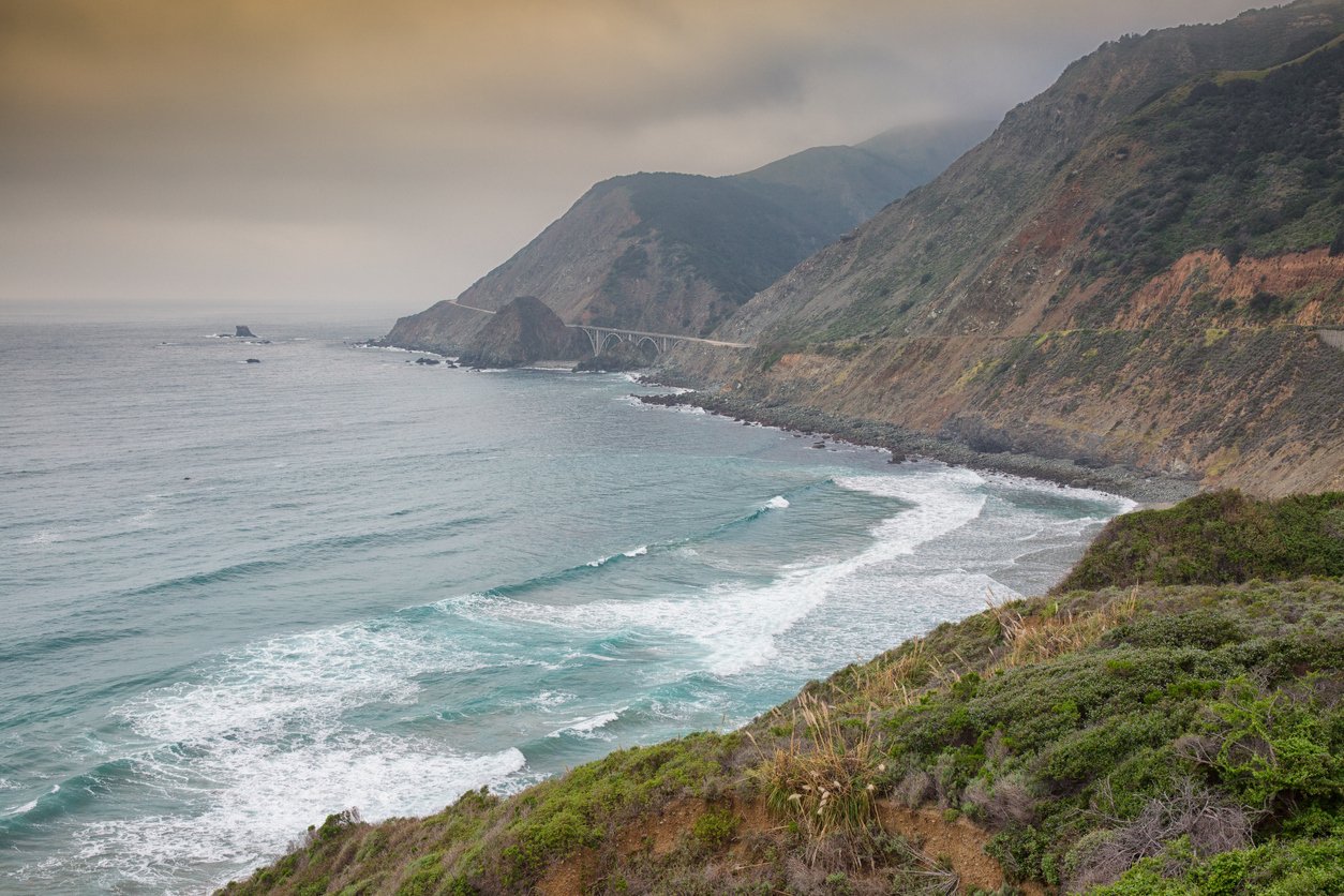 View of Big Sur’s steep coastline surrounded by mist during cloudy weather. The waves are pretty strong. There is a bridge in the distance.