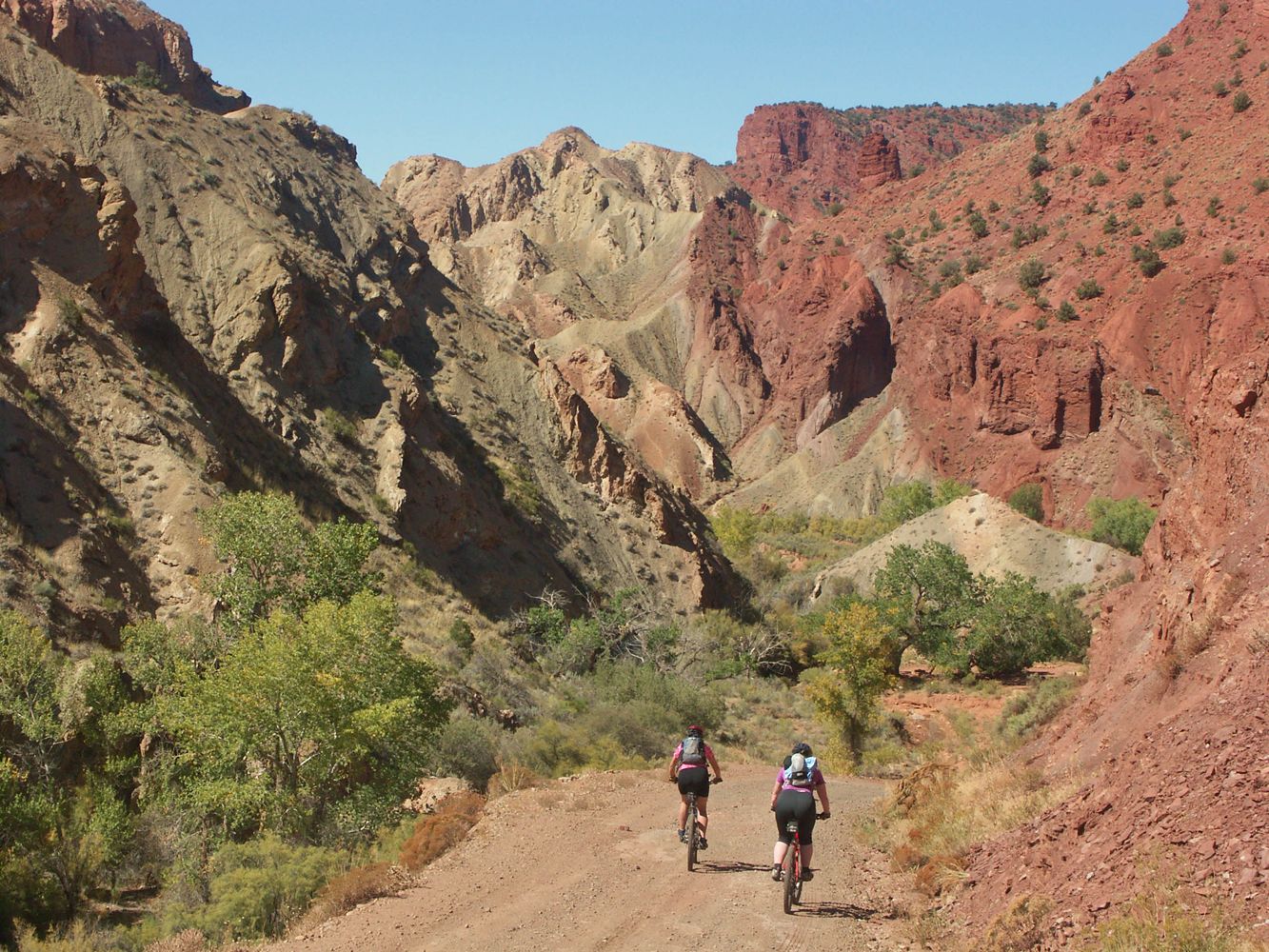 Mountain bikers ride a wide trail leading to colorful rock formations, along the Kokopelli Trail.