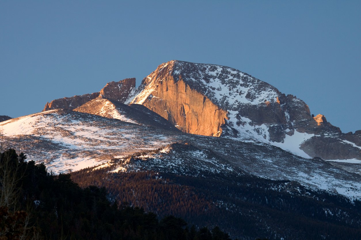 Morning sun light hits the east face or the sheer cliff known as "The Diamond" of the snow capped Longs Peak rising to 14,255 feet, in Rocky Mountain National Park, Colorado. Thick forest covers the lower elevations of the beautiful mountain.