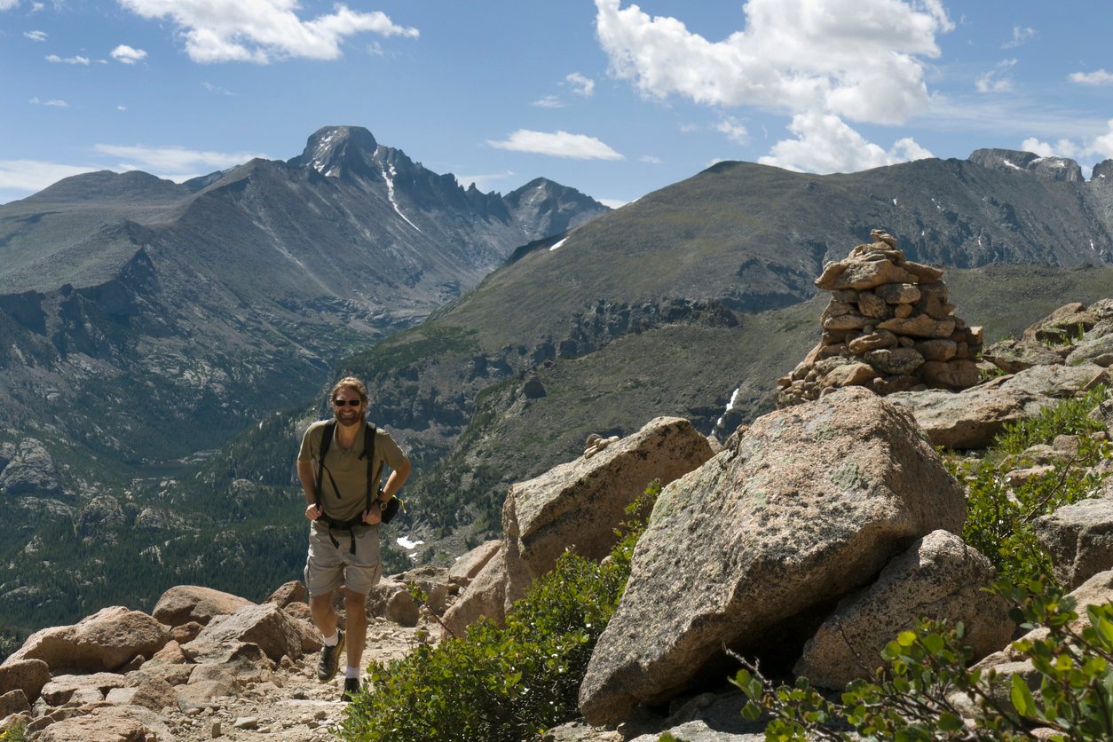 As clouds drift past the 14,255 foot Longs Peak and the rock formations known as the Keyboard of the Winds a bearded man with a backpack hikes up the Flattop mountains trail past a cairn and granite rocks in Rocky Mountain National Park, Colorado.