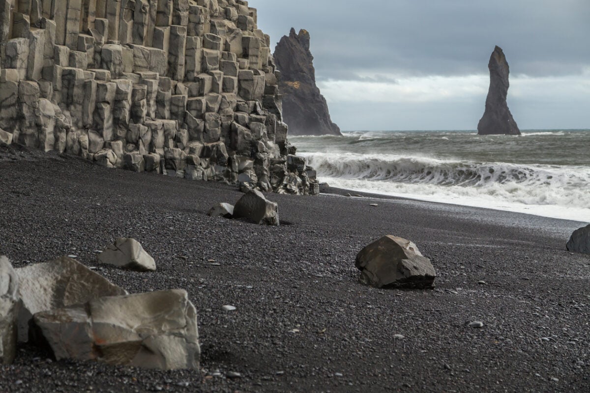 Basalt rock formations on the volcanic black sand beach at Vik in Iceland. The cliff face is columns of hexagonal rocks. Rock stacks are in the sea. Large waves and spray is being whipped up from the ocean. Rocks lie on the sand and the sky is cloudy and moody.