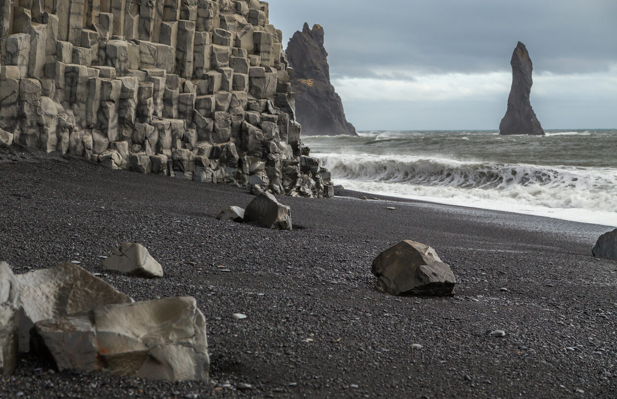 Basalt rock formations on the volcanic black sand beach at Vik in Iceland. The cliff face is columns of hexagonal rocks. Rock stacks are in the sea. Large waves and spray is being whipped up from the ocean. Rocks lie on the sand and the sky is cloudy and moody.
