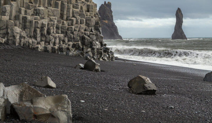 Basalt rock formations on the volcanic black sand beach at Vik in Iceland. The cliff face is columns of hexagonal rocks. Rock stacks are in the sea. Large waves and spray is being whipped up from the ocean. Rocks lie on the sand and the sky is cloudy and moody.