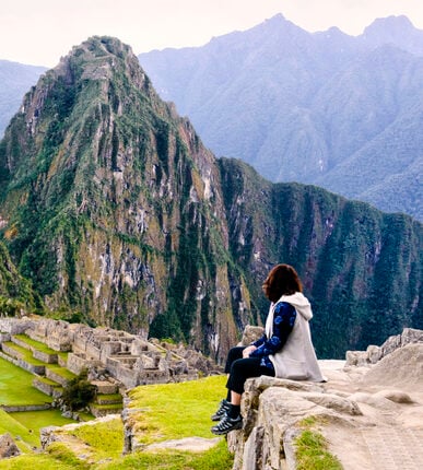 Woman sitting on a ledge and overlooking the Inca ruins of the city of Machu Picchu seen in the background.