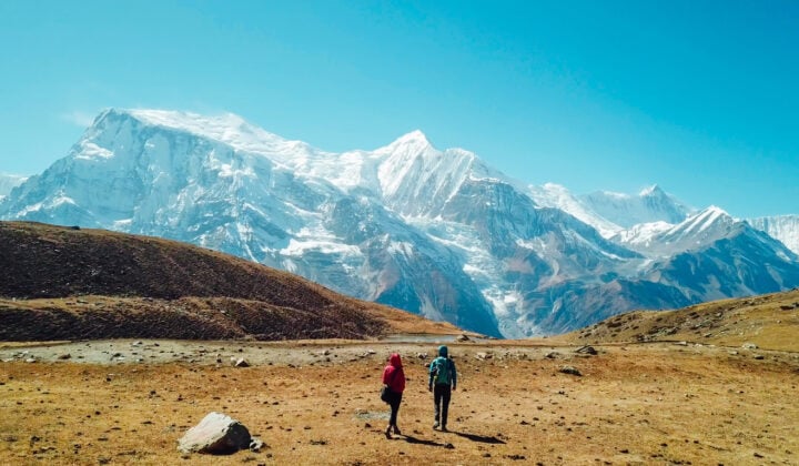 A couple walking towards the Ice lake, as part of the Annapurna Circuit Trek, Himalayas, Nepal. Annapurna chain in the back, covered with snow. Clear weather, dry grass, snowy peaks.