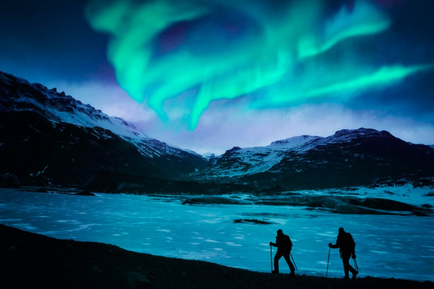 Winter hikers along a frozen lake and bathed in neon green northern lights