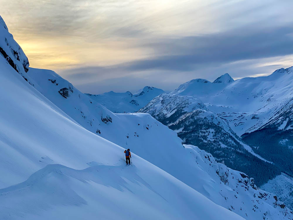 A backcountry skier moving horizontally across a steep powder-laden slope during twilight surrounded by peaks and valleys covered in snow-dusted conifers.