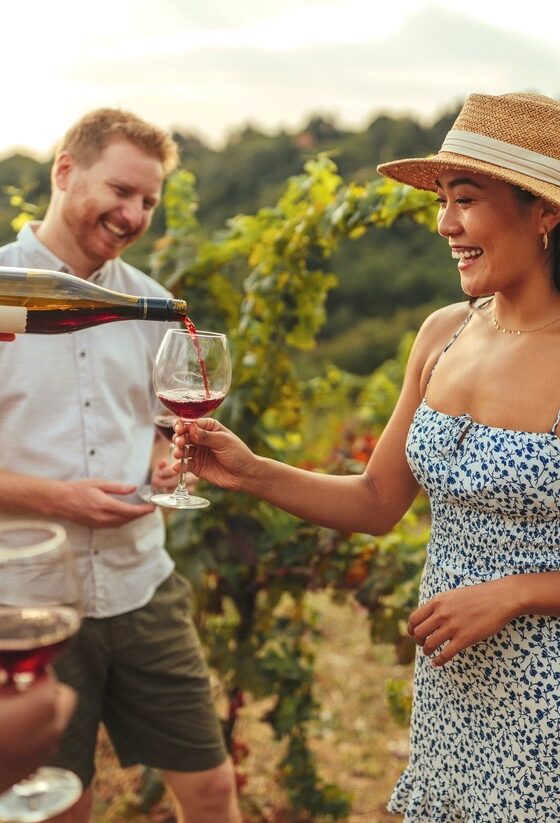 Unrecognizable man pouring red wine to asian woman in vineyard who came here with friends