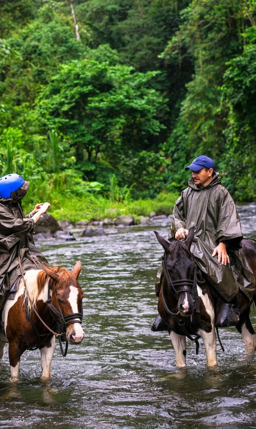 Woman taking selfie during Horseback riding on foggy and rainy day while crossing the river Arenal near small town La Fortuna in Costa Rica. Area is known as a gateway to Arenal Volcano National Park with two volcanoes, Active Arenal Volcano and Dormant Chato Volcano.