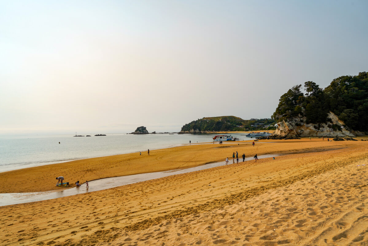 View of a sand beach with rock formations with trees in the background.
