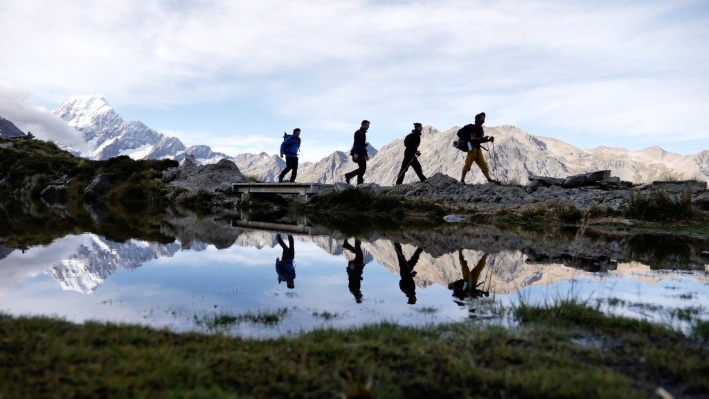 Four hikers walk in Mt Cook/Aoraki with snow-covered mountains in the background and a pool of water reflecting the scene in the foreground, 