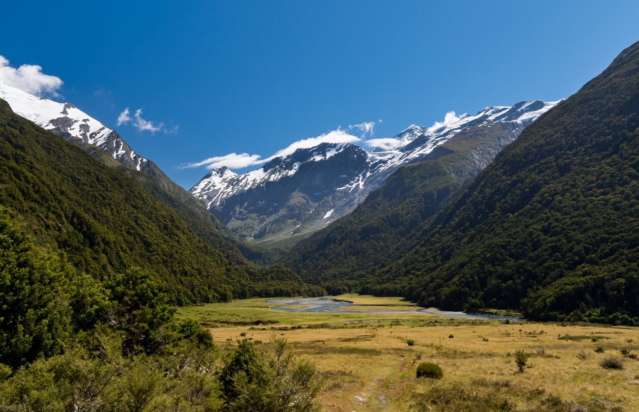 River valley surrounded by steep tree-covered mountains and one particularly high snow-covered mountain in the background.