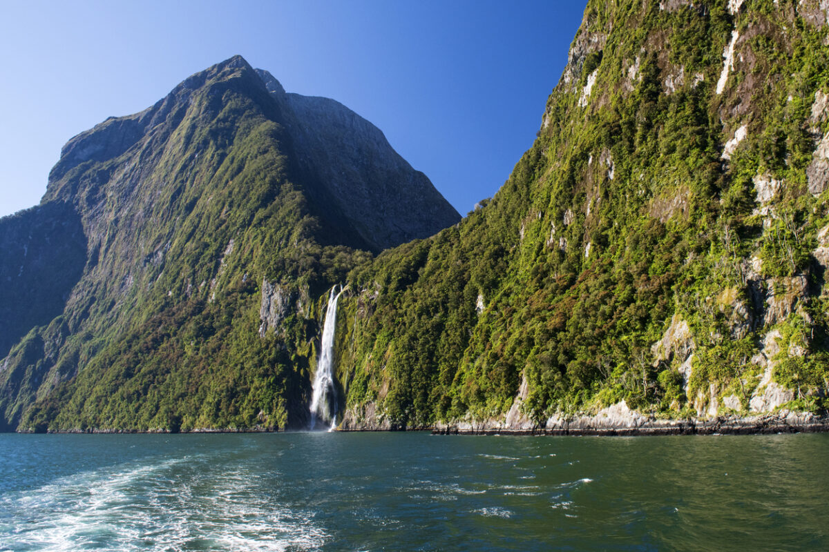 View of a tree-covered fjord wall from the water with a waterfall. 