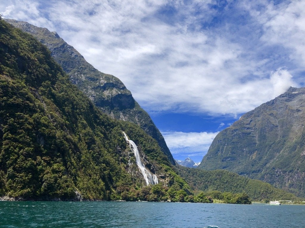 Sea-view of steep tree-covered cliffs and a waterfall with a snow-covered mountain peak in the distance.