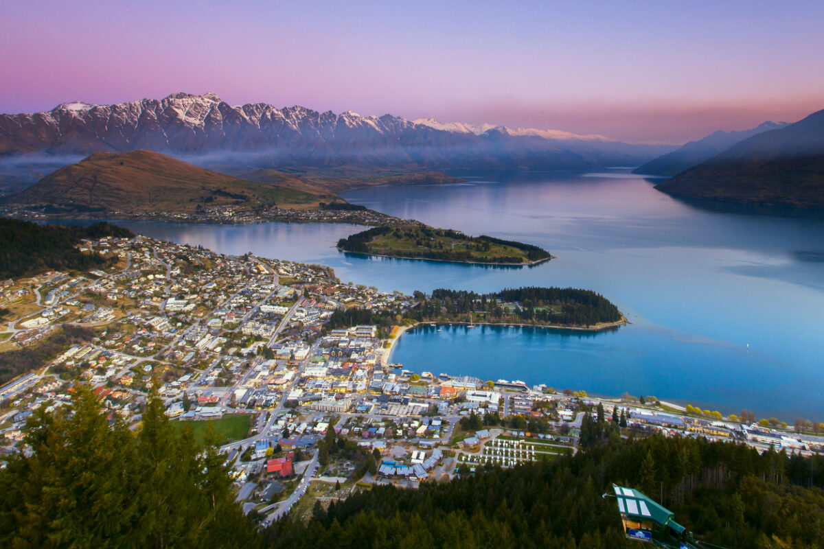 Aerial view of Queenstown during sunset with sea and steep snow-covered mountains in the distance. 