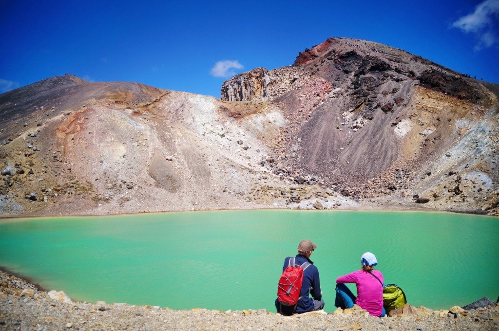 Two people sitting with a turquoise lake and a hill in front of them. 