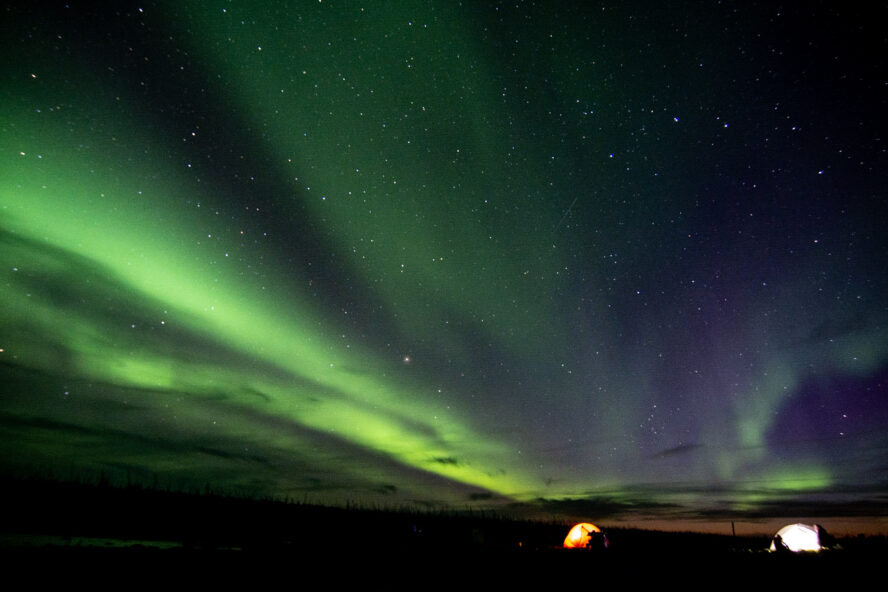 Neon green northern lights over two lit up tents alongside the Manitoba River in Canada.