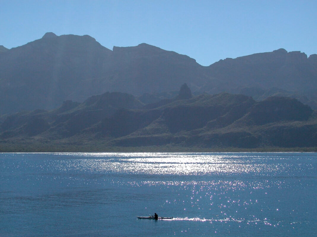 View of the Gigantia mountain range on the coast of the Baja California peninsula. In the front part of the picture there is a person kayaking, miniscule in size compared to the sea and the mountain. 