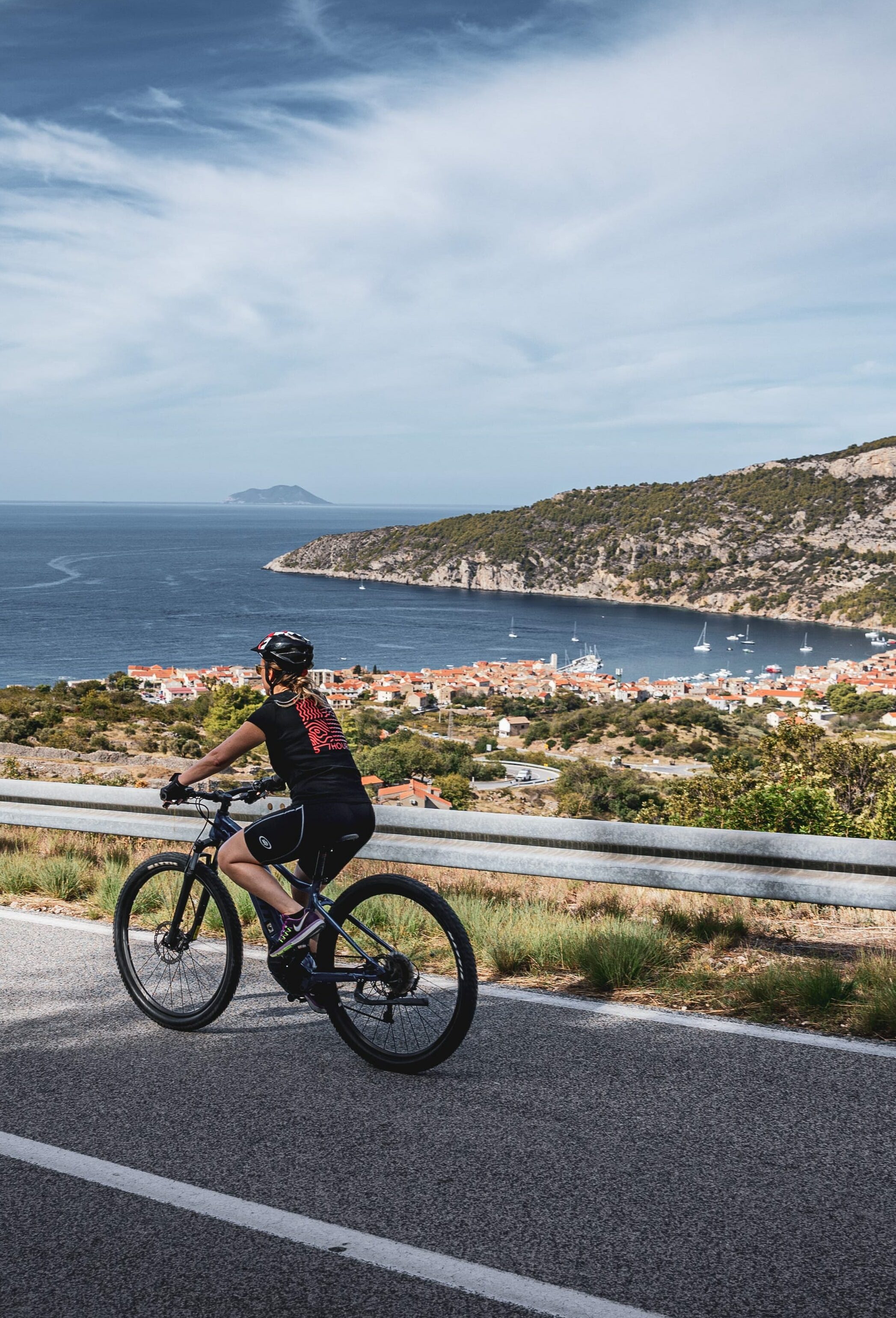 A person in a 57hours tshirt cycling along the coast on a road with a Dalmatian city and sea in the background