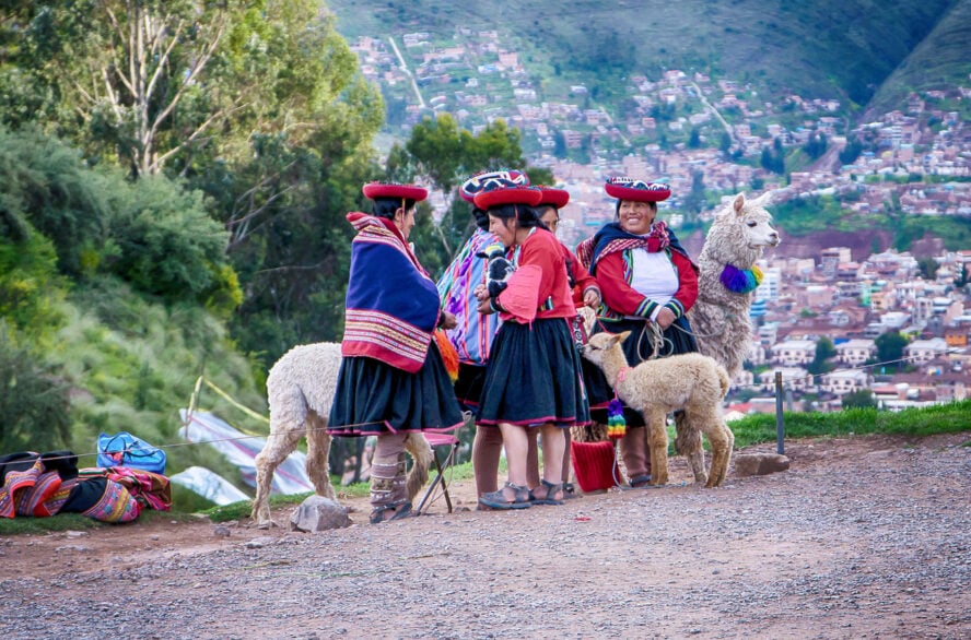 A group of women in traditional attire, alongside llamas with colorful scarves.