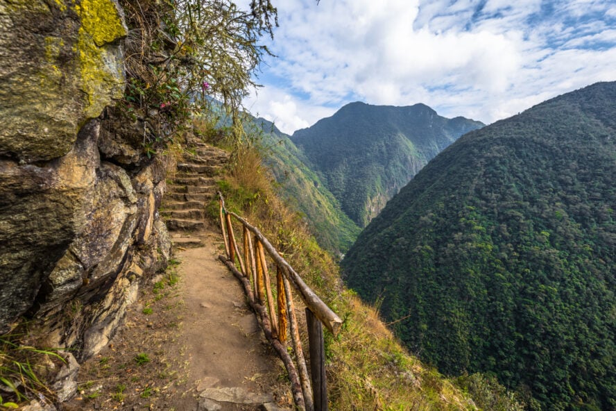 Dirt path and stone steps of the Inca Trail, running next to a steep cliffside, overlooking a lush green valley.