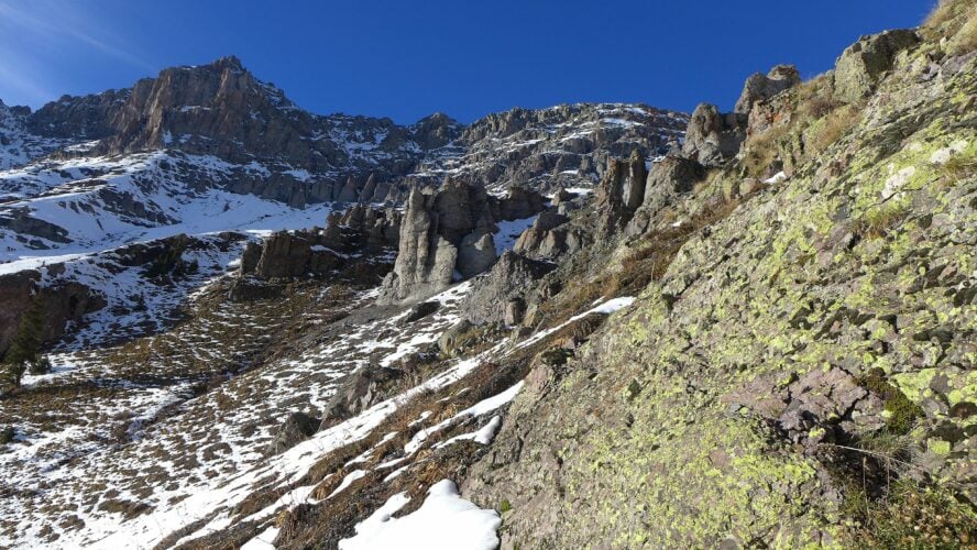 A rocky section of the trail on the Sneffels Highline loop showing gray mountain peaks, snow, and green lichen in the foreground. 