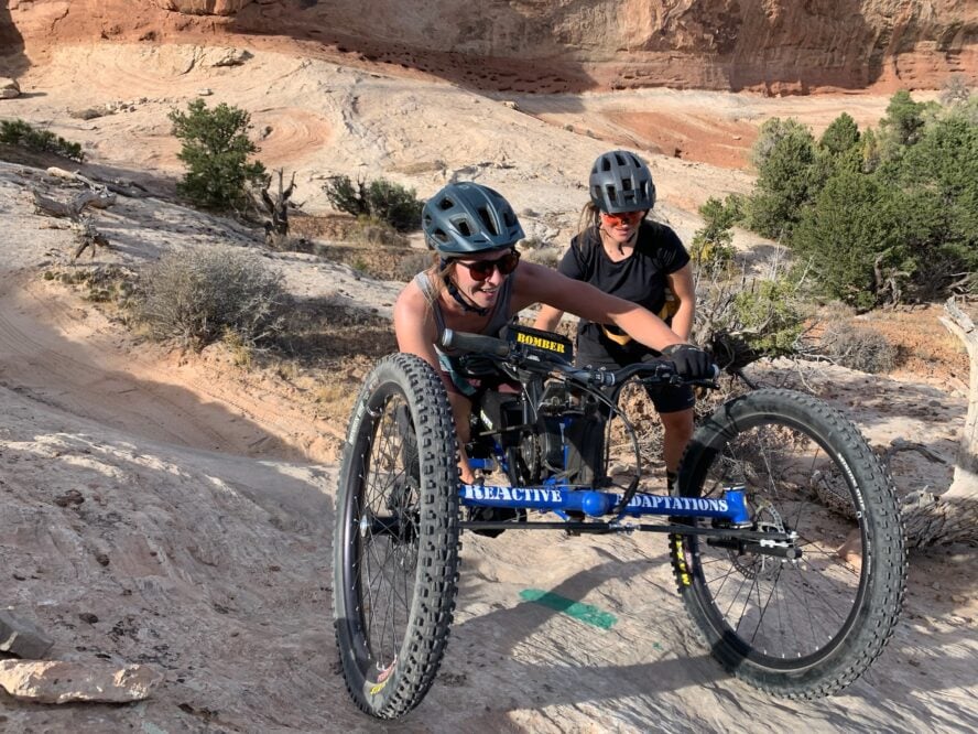 A mtb handcyclist ascending slick rock in Moab with a spotter behind.