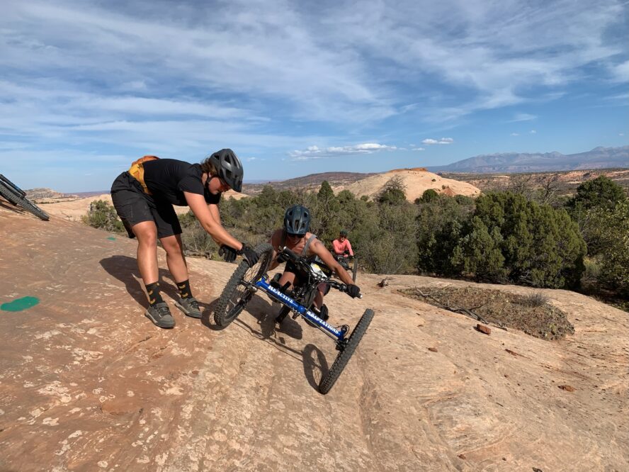 One handcyclist on a steep off-camber section of trail getting support so they don’t tip.