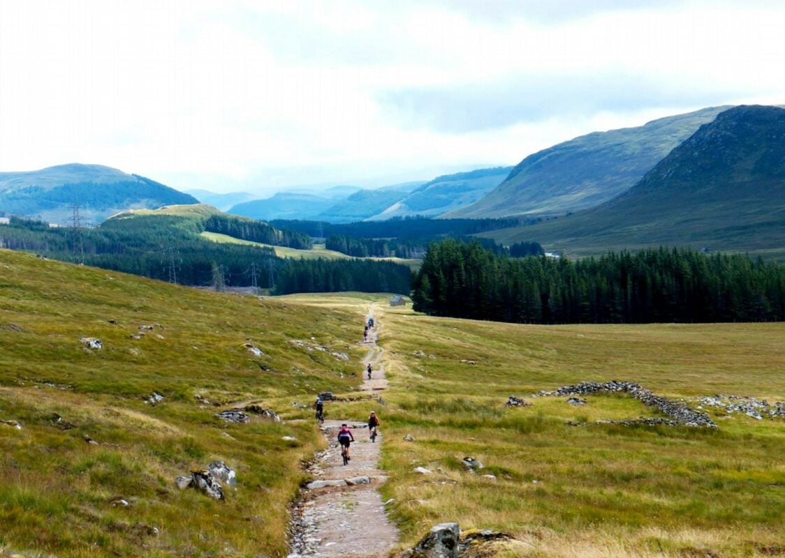 Mountain bikers riding a trail cutting a field in the Scottish Highlands surrounded by small conifer woods and low mountains. 