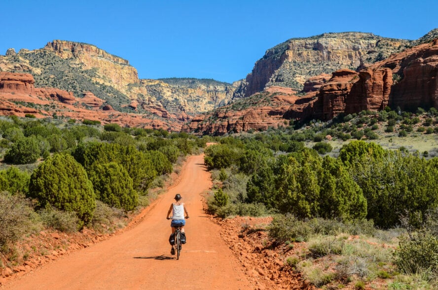 A biker riding on a smooth chaparral-lined trail towards massive sandstone formations