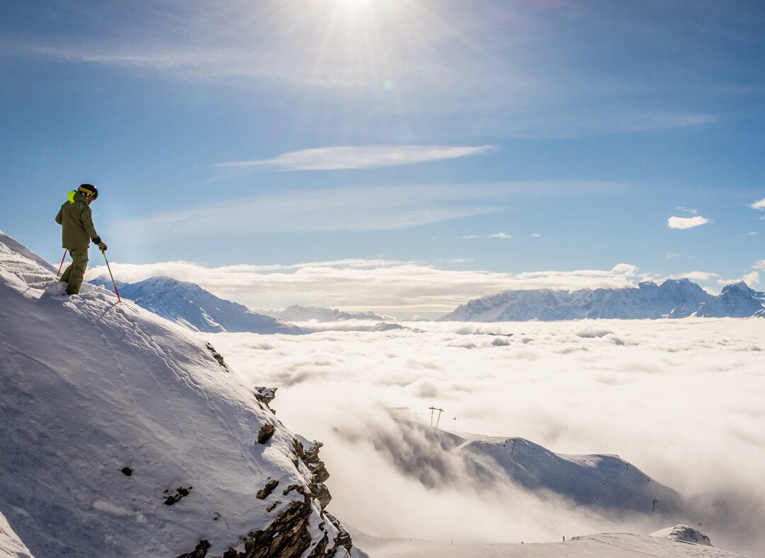 A skier in the Swiss ski resort of Verbier standing on top of a cliff above a sea of clouds, looking towards Mont Blanc and Chamonix.