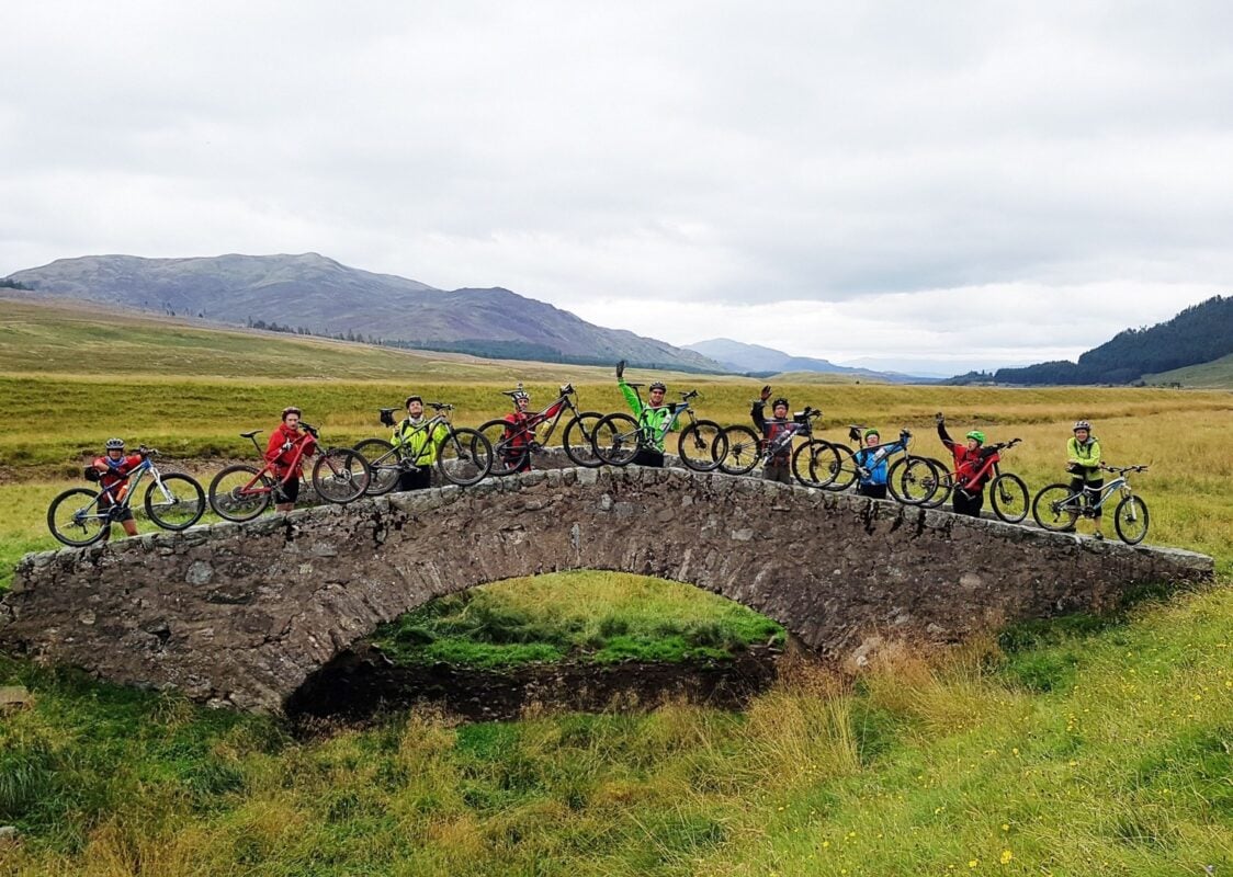 A group of mountain bikers posing for a photo on a stone bridge in the Scottish Highlands. 