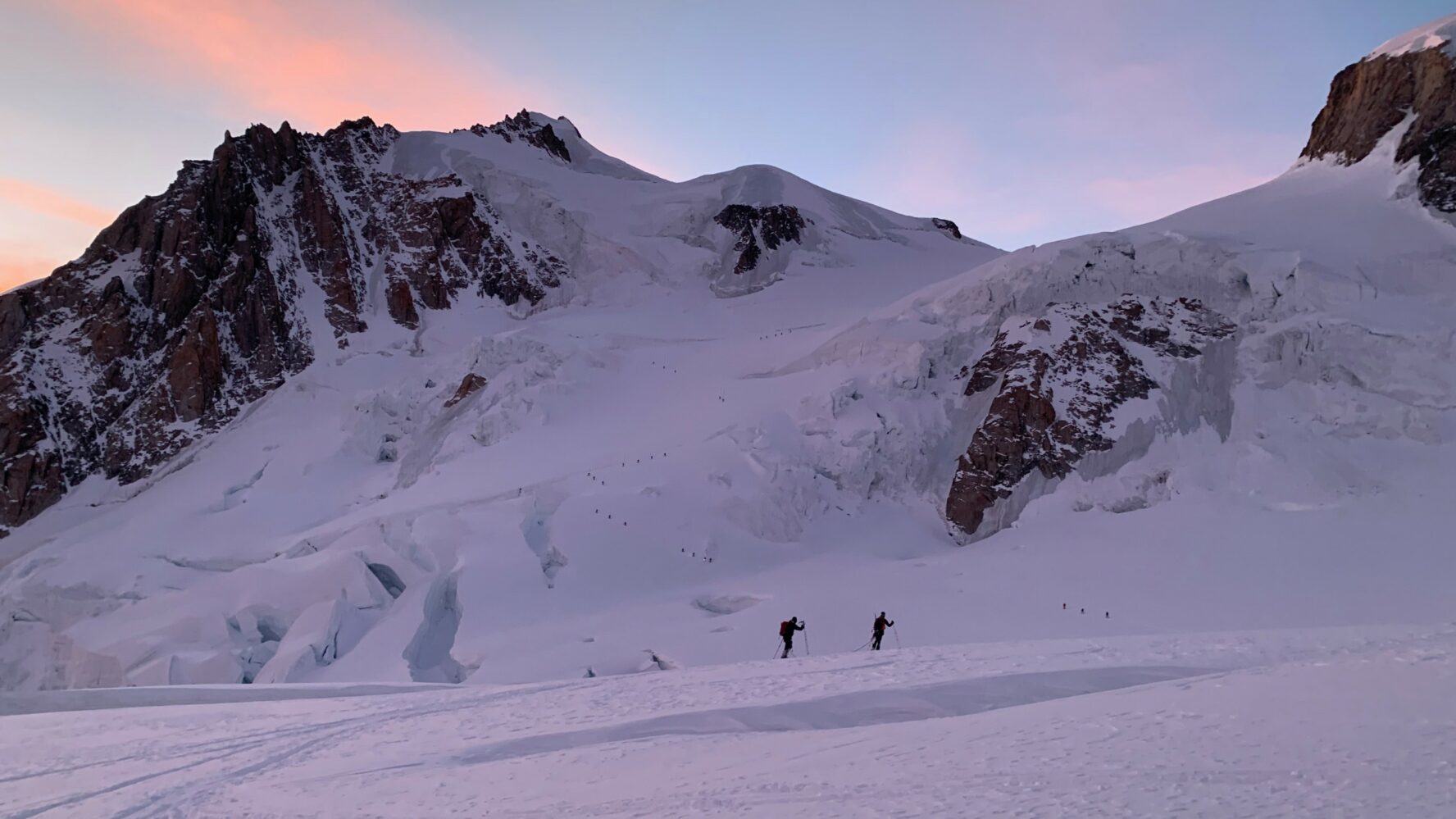 Two backcountry skiers skinning in the Alps among large peaks during sunset.