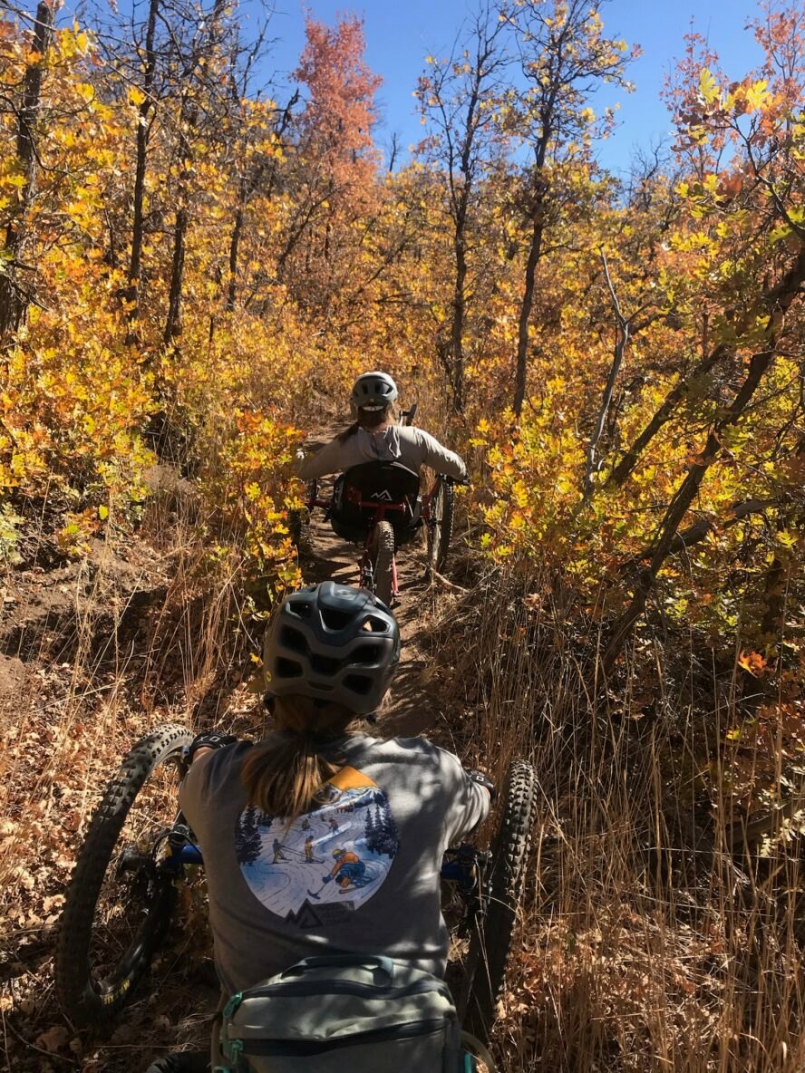 Two adaptive riders descending an overgrown trail, with grass at head height.