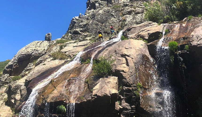 Two canyoneers reach the top of a rock formation on top of which flows a small waterfall using ropes.