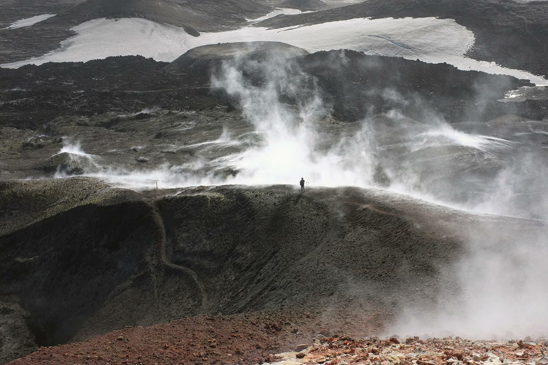 A hiker standing amidst s volcanic field shrouded in steam, photogrsphed by Jean-Christophe Bechet.