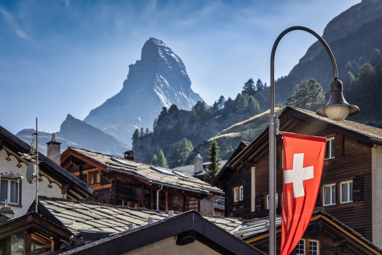 Zermatt Village Typical Traditional Swiss Alps Wooden Houses in the center of Zermatt Village with illuminated Swiss National Flag in the foreground. Famous Matterhorn Mountain Peak under blue summer sky in the background. Zermatt, Valais Canton, Switzerland, Europe