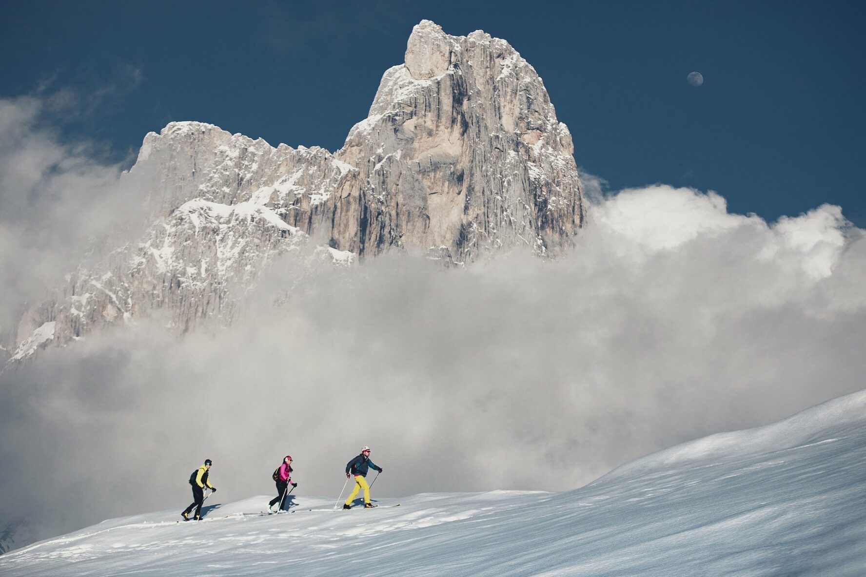 Three backcountry skiers skinning a moderate slope at very high altitudes around Ortler with a jagged peak in the background.