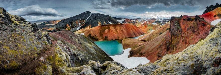 A panorama of the colorful rhyolite volcanic mountains of Landmannalaugar with a bright blue alpine lake in the center.