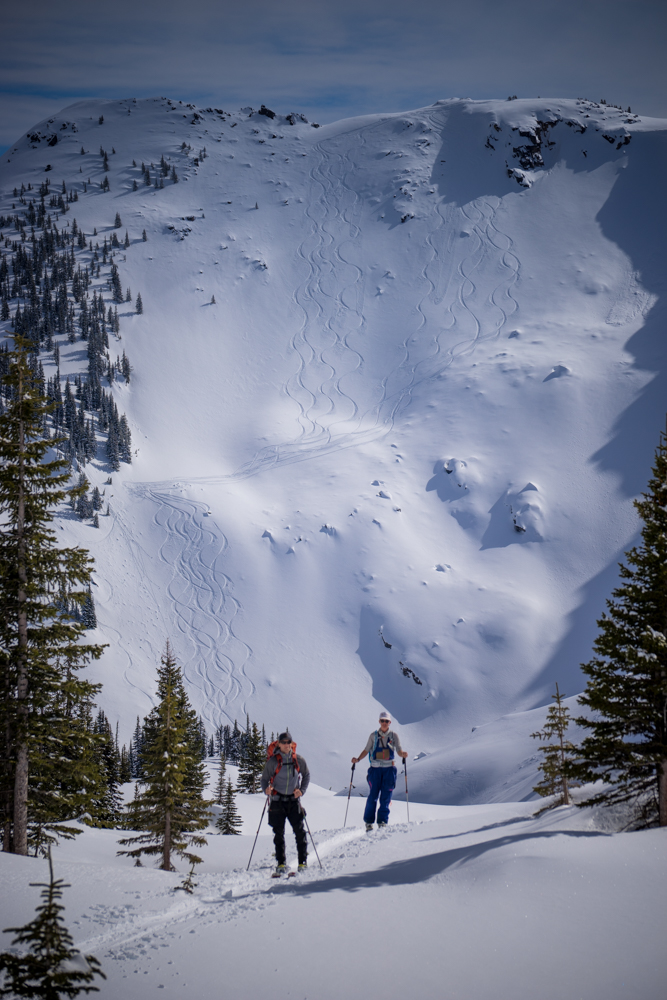 Two backcountry skiers at Mt Baker surrounded with conifers with very steep slopes tracked by skis behind them.