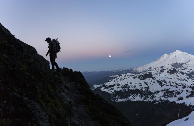 The silhouette of a climber scrambling up Shuksan’s Fisher Chimneys, as Mount Baker bathed in early morning light sits in the background.