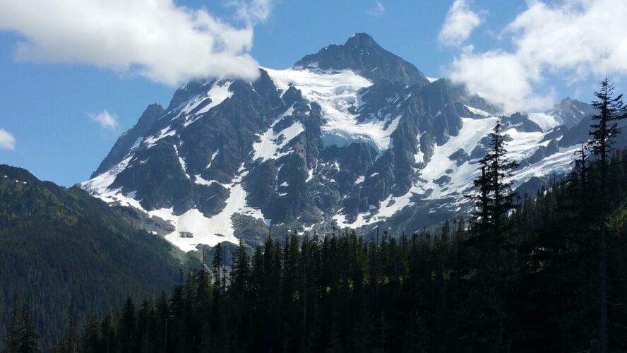 Mount Shuksan in Washington in the summer, full of glaciers and snow chutes, with forested hills in front.