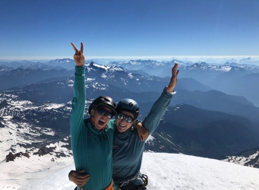 Guides Avery Solte (left) and Victoria Garvin (right) stand on the summit of Mount Baker with the cascades extending seemingly infinitely in the background.