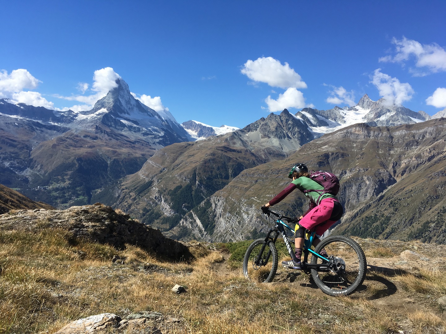 A woman MTB rider is riding a trail near Verbier in the Swiss Alps above treeline surrounded by views of lofty peaks.
