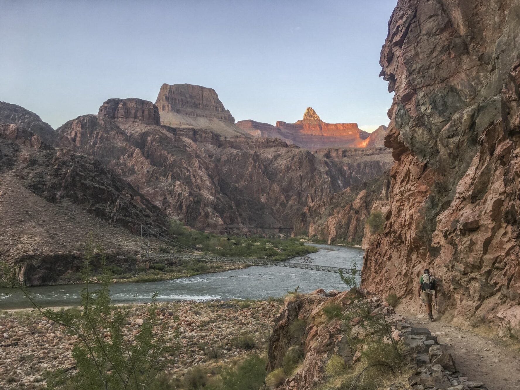 A hiker nearing a bridge over a Grand Canyon river with a vista in the background.