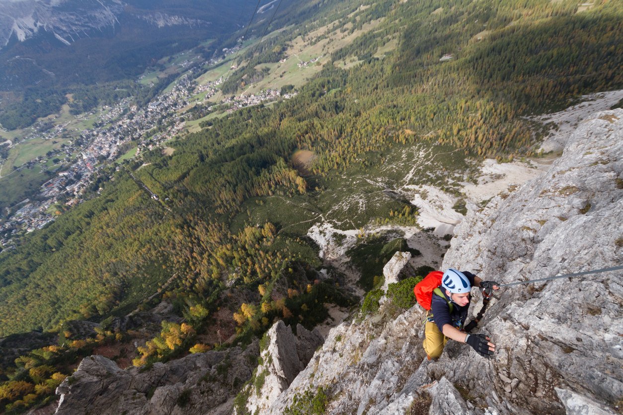 A rock climber tackling a climbing route in the Dolomites with views of a forest and Cortina d’Ampezzo in the distance.