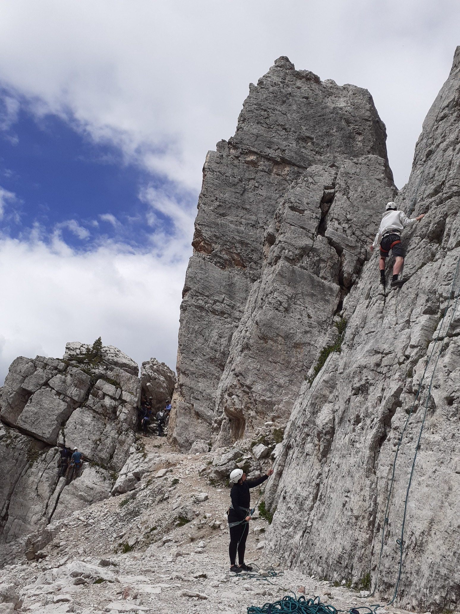 A climber tackling a route and another climber helping while standing at the foot of a creviced limestone wall in the Dolomites.