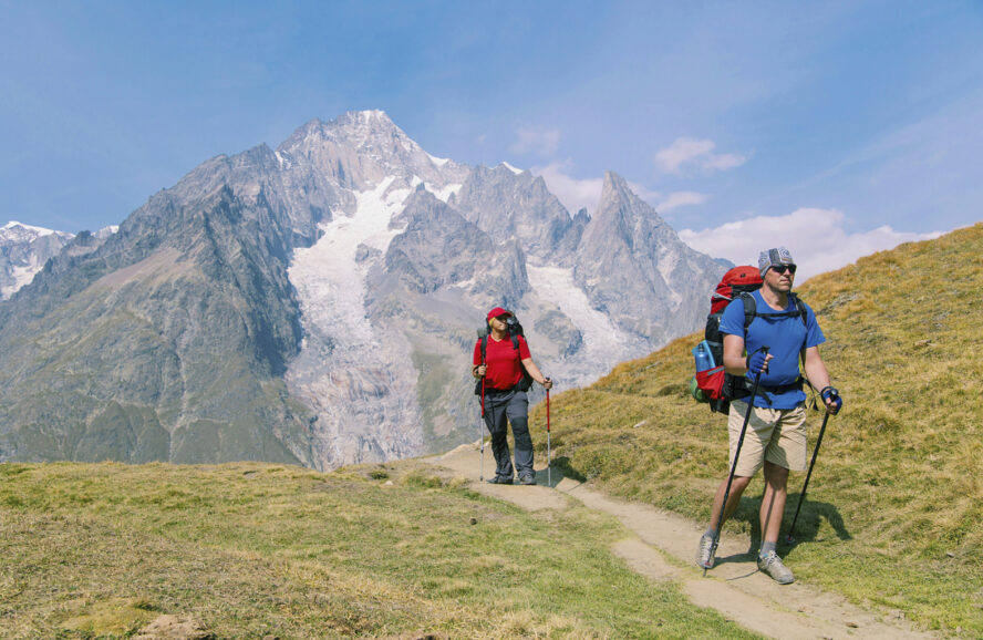 Two hikers walking a trail along the famous Tour du Mont Blanc with views of lofty snow-capped peaks behind them.