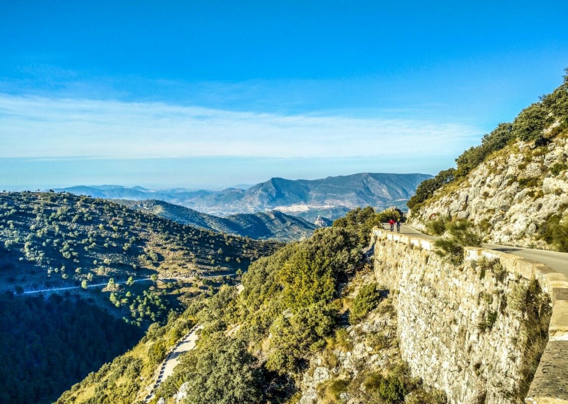 Two cyclists riding along a road in the Los Alcornocales Natural Park, characterized by rugged hilly Mediterranean landscapes.